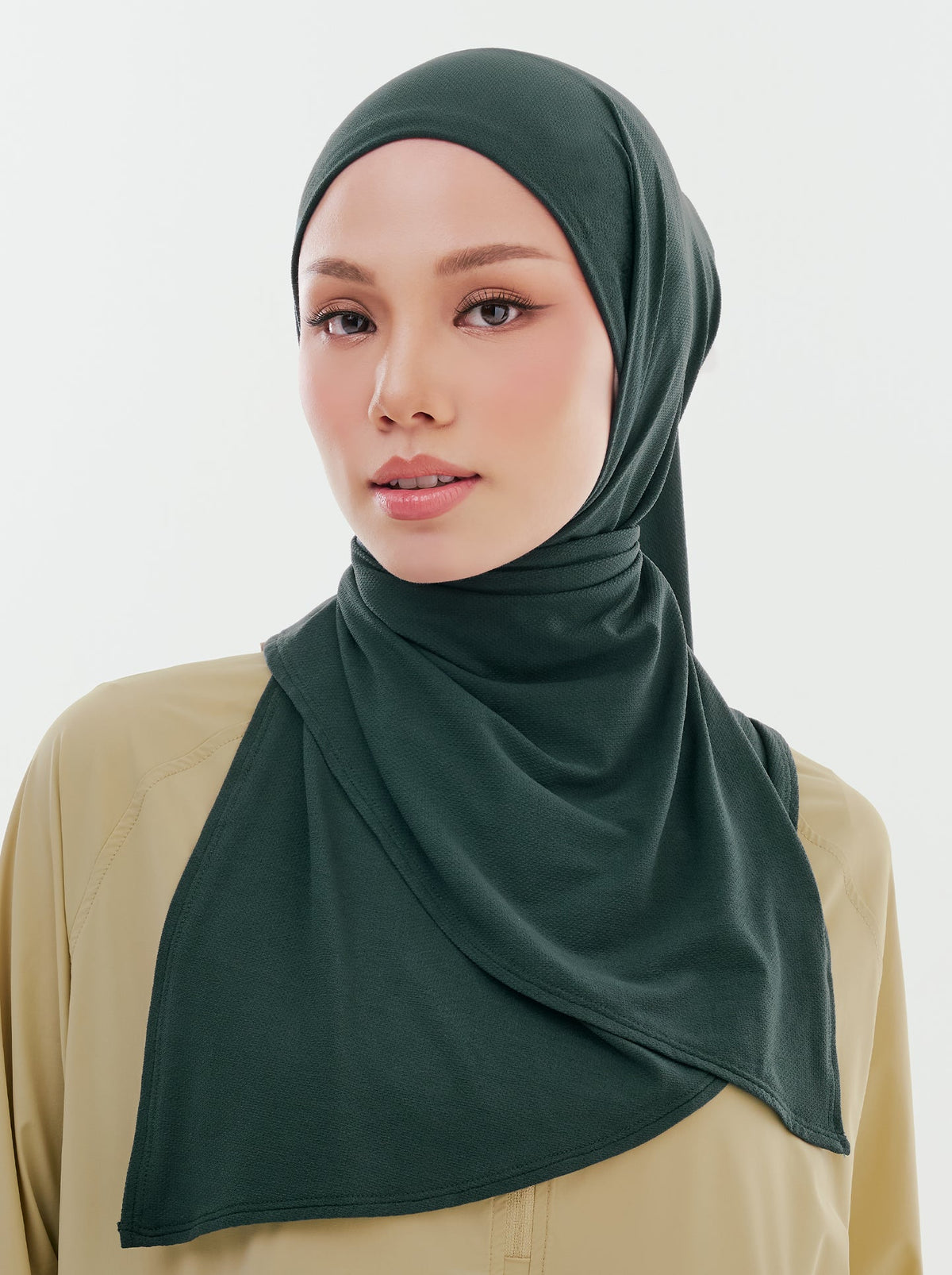 A woman in a dark green Najwa Sport hijab and olive top, standing relaxed against a plain background