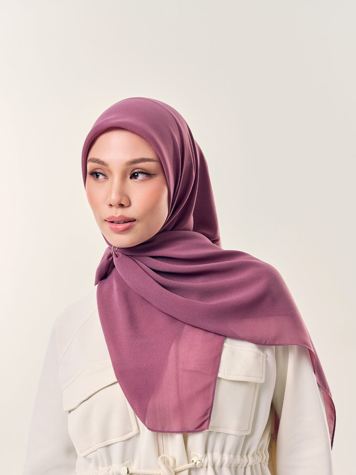 A woman in a mauve chiffon square tudung fastened neatly with a small pin under the chin, worn with a cream utility jacket