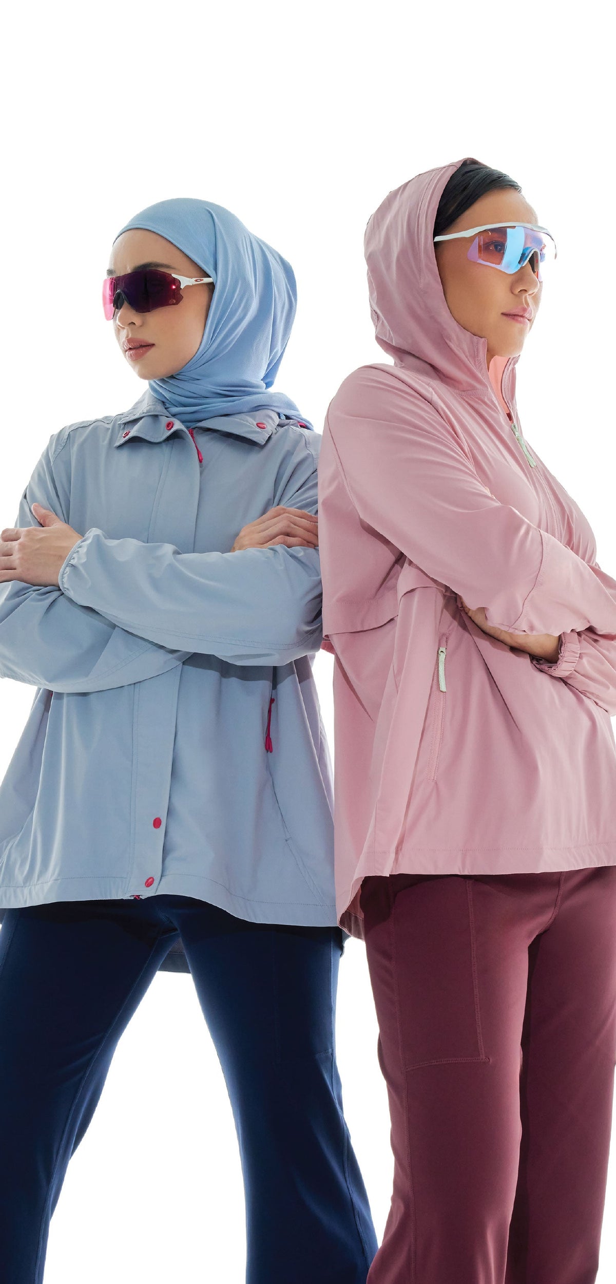 Two women in athleisure parkas and sports sunglasses standing back to back, one in a light blue hijab and one with a pink hood up