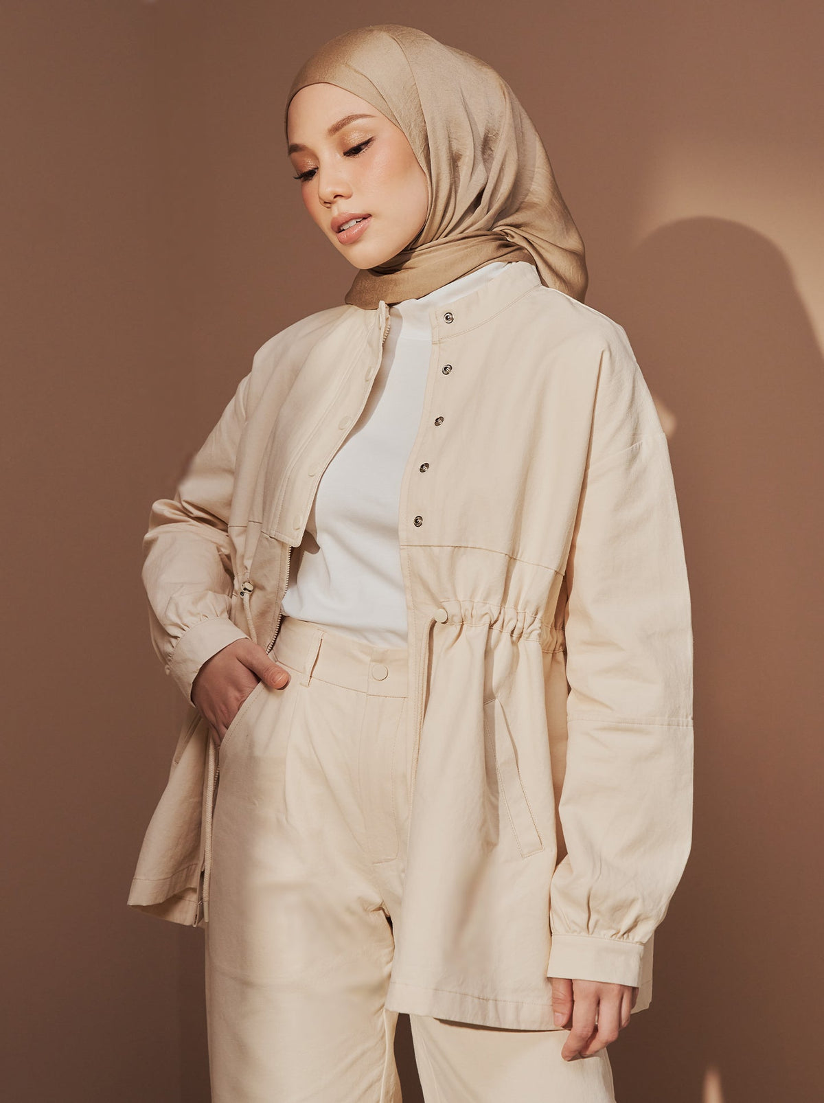 A woman in a golden beige satin square hijab, a cream field jacket, a white top and cream trousers, in a relaxed travel outfit