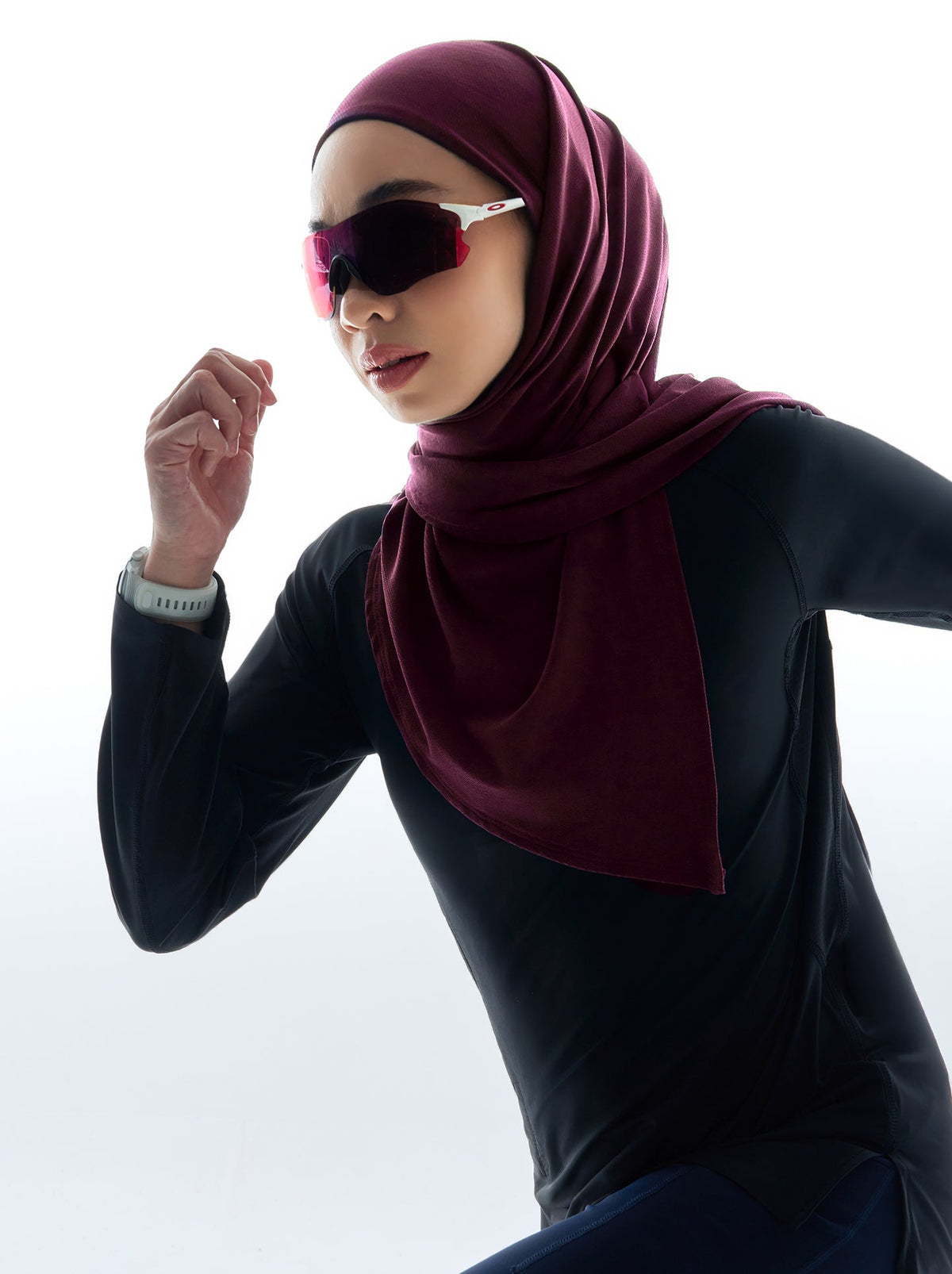 A runner in a burgundy sports hijab, black long-sleeved top and sports sunglasses, mid-stride against a white background