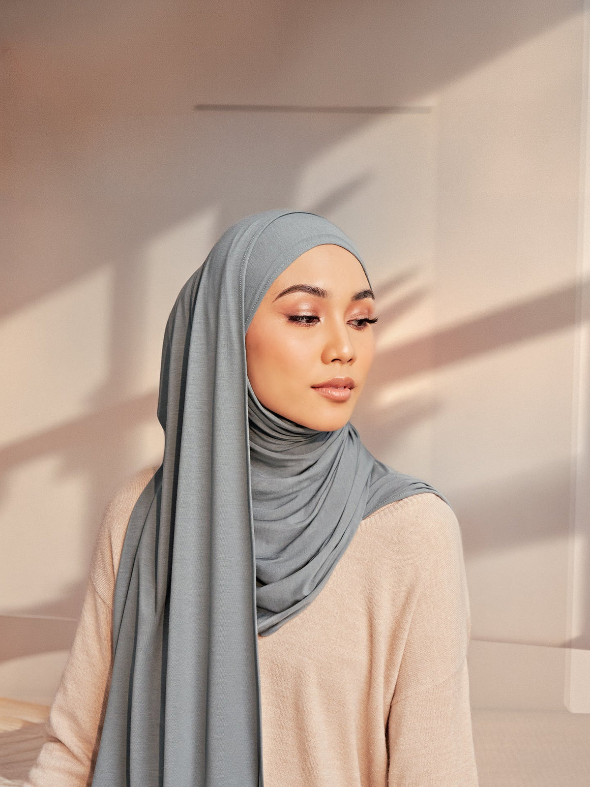 A woman in a grey-blue bamboo jersey shawl wrapped softly at the neck, one long end falling down the front over a beige knit jumper