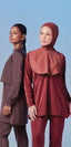 Two women in TudungPeople Flow and Wave swimwear sets, one in a mauve long-sleeve tunic and one in a rust-red swim hijab with cape, against a blue backdrop.